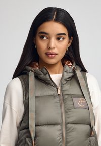 Wype�łniona oliwkowa zielona kamizelka z kapturem, z brązowymi akcentami i detalem logo "POLO CLUB". Zapięcie na zamek błyskawiczny z przodu i teksturowana tkanina.