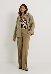 Mujer vestida con pantalones de pierna ancha beige, blusa estampada, chaqueta beige texturizada y zapatos cerrados beige con ojales metálicos, de pie sobre un suelo blanco.