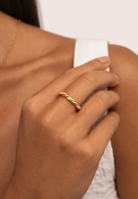 Close-up van een hand met een getinte huid die een gedraaide gouden ring met kleine diamanten op de ringvinger draagt, tegen een witte mouwloze top.