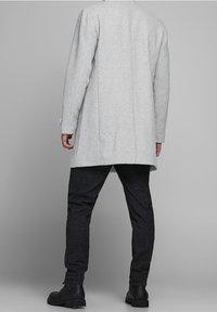 Mann trägt hellgraßen langen Mantel, dunkel karierten Hose und schwarze Schnürstiefel, steht mit dem Rücken zum grauen Hintergrund.