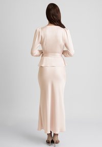 Jasnoróżowa satynowa sukienka z długimi bufkami, w talii peplum oraz dopasowaną spódnicą. Gładka faktura, długość midi, w połączeniu z beżowymi szpilkami.