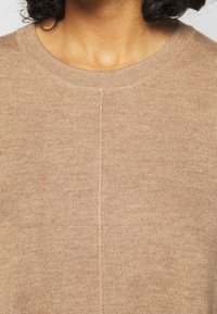 Pull en tricot beige avec un col rond, présentant une texture fine et des détails de couture subtils à l'avant et à l'arrière.