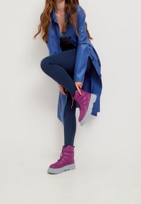 Botines acolchados morados con suela gris, combinados con un abrigo azul y leggings de color marino. Las botas cuentan con una cremallera lateral y cordones.