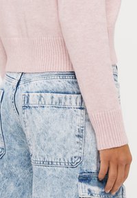 Jasnoróżowy sweter z dzianiny z prążkowanym wykończeniem na dole, połączony z wyblakłymi niebieskimi spodniami typu cargo z dużymi bocznymi kieszeniami i luźnym krojem.