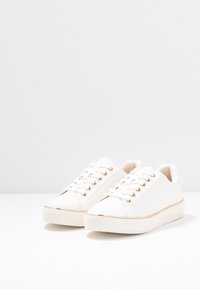 Baskets blanches avec une texture lisse, bout rond et œillets dorés. Présentent une silhouette basse et une semelle plateforme légèrement surélevée.