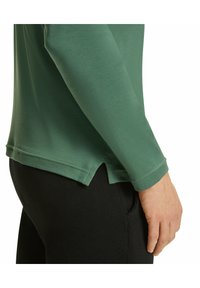 Grünes Langarmshirt mit abgerundetem Saum und seitlichen Schlitzen, kombiniert mit schwarzen, engen Hosen. Der Stoff sieht glatt und elastisch aus.