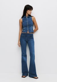 Denim cropped top met een ritssluiting en kraag, gecombineerd met uitlopende denim broeken. Beide zijn in een medium blauwe tint met zichtbare naaddetails.