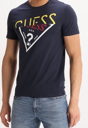 T-shirt en coton bleu marine avec un grand graphisme triangulaire blanc, du texte rouge et jaune, et le logo "GUESS" affiché en évidence.