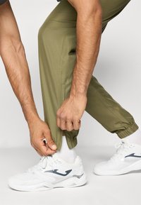 Olivengrønne treningsbukser med elastiske mansjetter, teksturert stoff, sidelommer, og en person som justerer leghemmen med hvite joggesko og sokker.