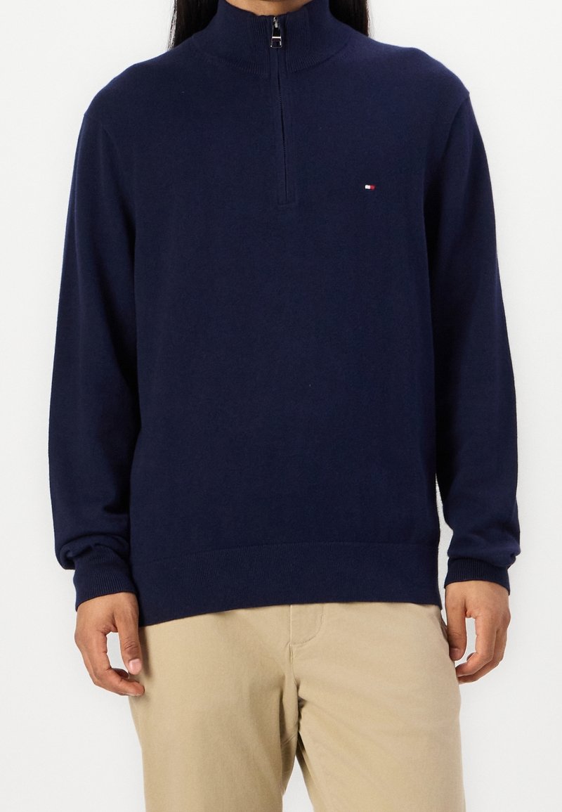 Personne portant un pull à manche longue bleu marine avec une fermeture éclair à mi-chemin et un petit logo sur la poitrine gauche, assorti à un pantalon beige sur un fond blanc uni.