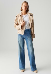 Manteau trench beige avec larges revers, boutons double boutonnage, associé à un jean à jambes larges bleu clair avec coutures avant et poches.