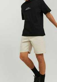 Osoba oblečená v černém tričku s bílým textem, béžových šortkách, černých ponožkách a černých teniskách, stojící na jednobarevném světlém pozadí.