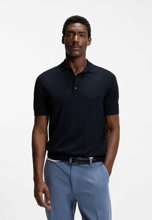 Uomo in piedi con le mani in tasca, indossa una polo nera a maniche corte e pantaloni azzurri chiaro su sfondo bianco.