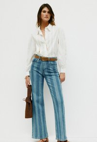 Hvid, langærmet skjorte med hulmønster; blå stribede flared jeans; brun bælte; tekstureret brun taske i hånden; sandaler.