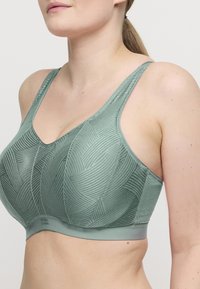 Mujer vistiendo un sujetador inalámbrico verde con estampado y tirantes anchos, mostrando el escote y la parte superior del torso contra un fondo simple.