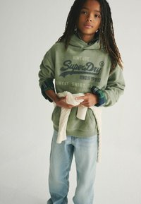 Sudadera verde con el logotipo "SuperDry", jeans de corte relajado en azul claro y un suéter de punto color crema atado a la cintura. La modelo tiene el cabello largo y texturizado.