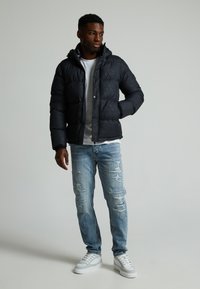 Chaqueta de plumas negra con capucha, con un patrón texturizado, cierre de cremallera, combinada con jeans desgastados de color azul claro y zapatillas blancas.