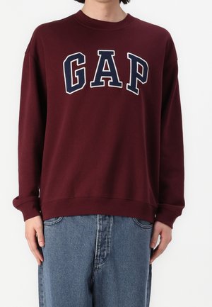 Person trägt einen bordeauxfarbenen Pullover mit "GAP" in großen marineblauen und weißen Buchstaben, kombiniert mit lockeren blauen Jeans.