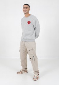 Grauer Sweatshirt mit einem roten Herz-Logo auf der linken Brust, kombiniert mit beigen Cargo-Hosen, die mehrere Taschen und Reißverschlüsse haben. Braune Sneakers.