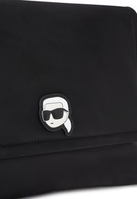 Borsa nera in nylon con un prominente emblema in gomma raffigurante un volto con occhiali da sole e una coda di cavallo. Design semplice con una superficie liscia.
