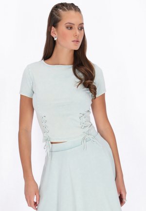 Femeie cu părul împletit, purtând un top bleu cu mâneci scurte și șireturi laterale și o fustă asortată, uitându-se spre stânga.