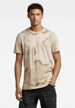 T-shirt in cotone beige con un motivo mimetico in tonalità più chiare, colletto rotondo e maniche corte. Tessuto lavabile in lavatrice.