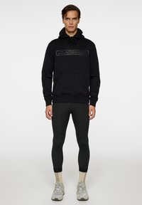 Mustat hupparit, joissa on teksturoitu logo, kengurutasku; yhdistetty mustiin legginsseihin ja vaaleanharmaisiin kengät, joissa on beige yksityiskohtia.