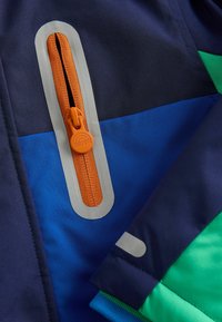 Jūros laivyno ir mėlyna medžiaga su žaliais akcentais, pasižymi oranžine užtrauktuku su apvaliu traukimo elementu ir kontrastinga pilka užtrauktuko apdaila.