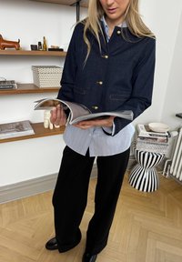Veste en denim avec des boutons dorés, de couleur marine, associée à une chemise rayée bleu clair et un pantalon large noir. Le mannequin tient un magazine ouvert.