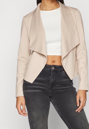 Kvinde iført en beige jakke med åben front over en hvid cropped top kombineret med sort højtaljede jeans, stående med armene afslappede.