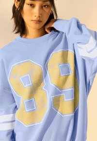 Felpa oversize blu chiaro con grandi numeri gialli "89" sul davanti, strisce bianche sulle maniche e una texture morbida.