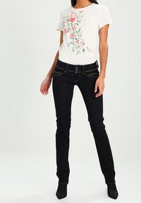 Kvinna klädd i vit kortärmad t-shirt med rosa blommönster och slim-fit mörkblå jeans, står med ena handen hängande ner och den andra något upphöjd.