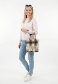 Mujer sonriente con suéter rosa claro y jeans azules, sostiene un bolso tote marrón a cuadros con flecos, lleva zapatillas blancas y reloj de pulsera morado.