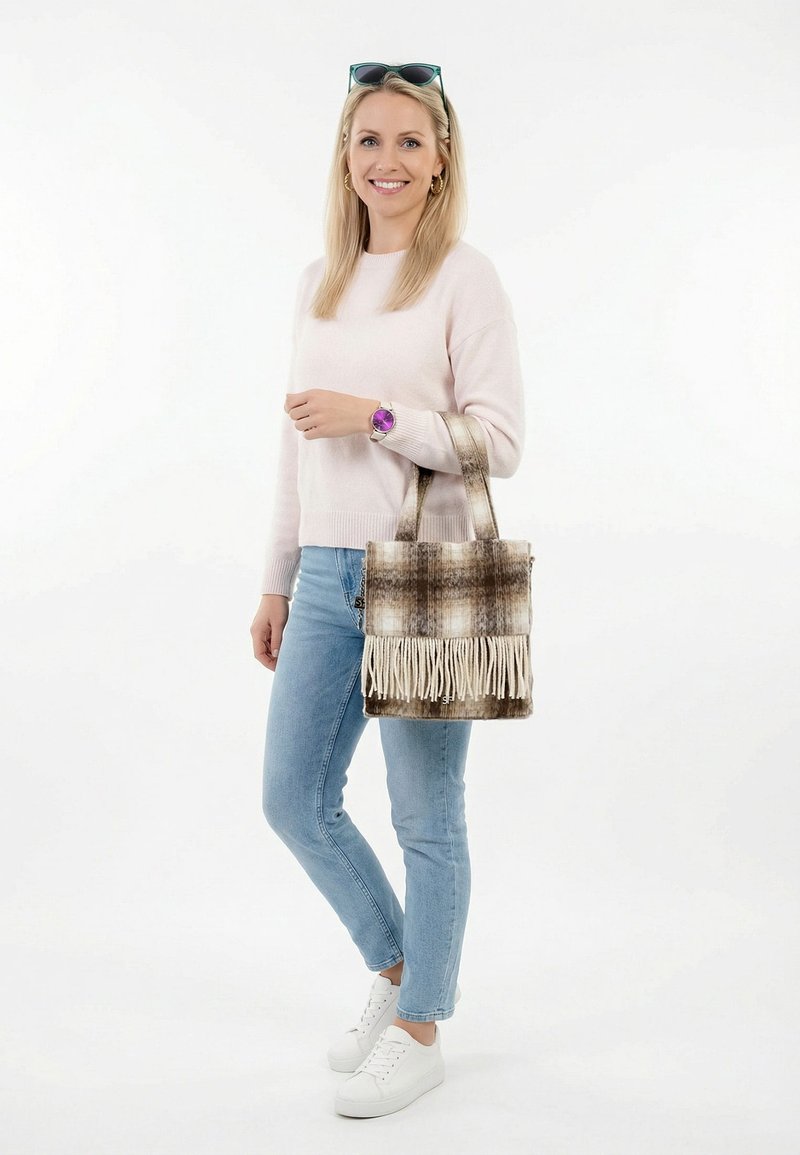 Mujer sonriente con suéter rosa claro y jeans azules, sostiene un bolso tote marrón a cuadros con flecos, lleva zapatillas blancas y reloj de pulsera morado.