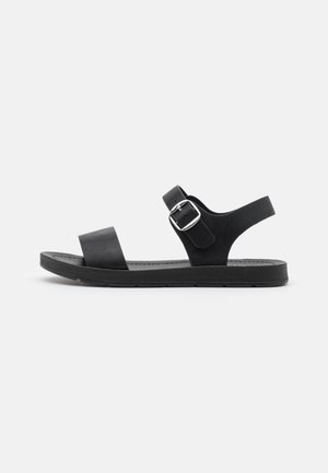 Black flat sandal with a single front strap, ankle strap with silver buckle, and cushioned sole on a white background.