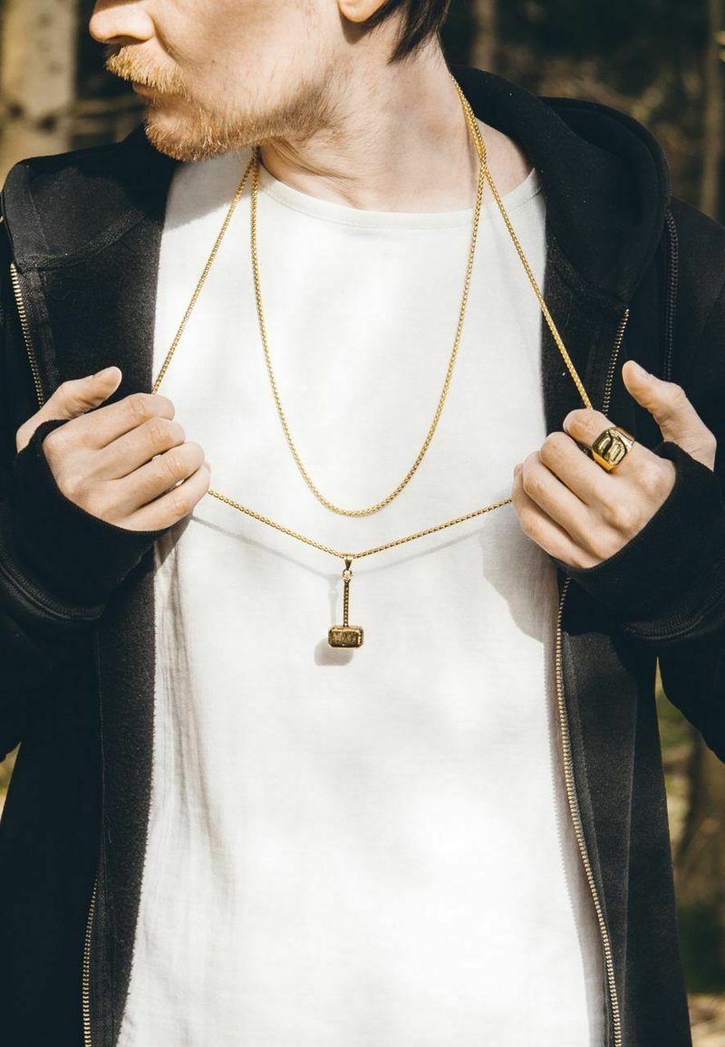 Gold-tone jewelry includes a long necklace with a hammer pendant and a ring, worn over a white shirt and black hoodie.