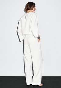 Weißer Leinen-Jumpsuit mit langen Ärmeln und weitem Bein. Mit einem Rückenausschnitt und lockerem Sitz. Kombiniert mit schwarzen High Heels.