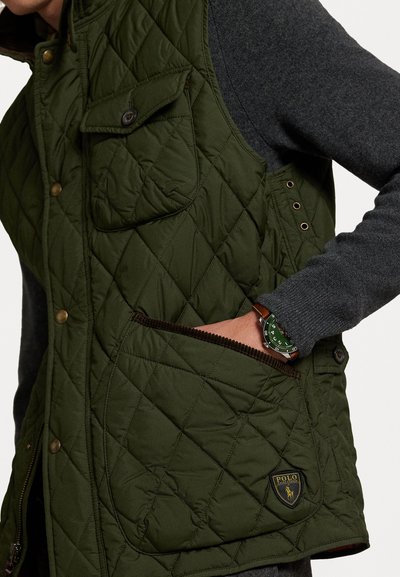 Zaļa izšūta veste ar priekšējām kabatām, spiedpogām un ribotām akcentiem. Iespējojama ar logo uzplecu un kontrastējošu pelēku garām piedurknēm kreklu zemāk.