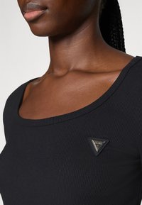 Close-up of a person wearing a black ribbed top with a small triangular Guess logo badge near the collarbone.