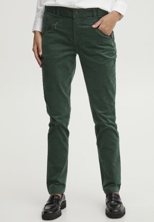 Pantaloni in velluto a coste verde con un design aderente, tasche frontali con zip e una texture liscia. Indossati con mocassini neri.