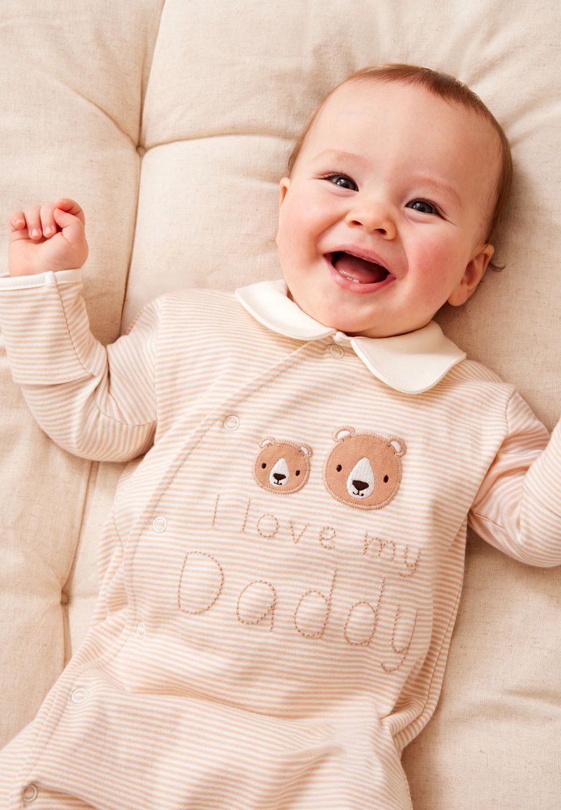 Bébé souriant en grenouillère beige rayée avec des visages d'ours et le texte « J'aime mon papa », allongé sur une surface beige rembourrée.
