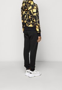 Haut à capuche noir avec un motif de chaînes dorées, joggings noirs et baskets blanches avec des accents argentés. Design détaillé avec une texture distincte.