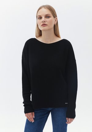 Schwarzer gestrickter Pullover mit weitem Bootsausschnitt, langen Ärmeln und gerippten Bündchen. Kombiniert mit blauen Jeans, vor einem weißen Hintergrund präsentiert.