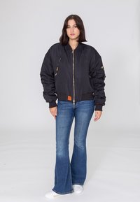 Czarna kurtka bombowca z zamykanymi kieszeniami, ribbowanymi mankietami i dolną krawędzią. Połączona z rozszerzającymi się niebieskimi dżinsami i białymi trampkami, na jednolitym tle.