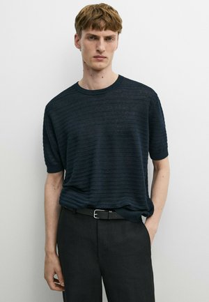 Jeune homme aux cheveux courts et bruns, portant un pull texturé bleu marine foncé à manches courtes et un pantalon noir avec une ceinture noire.