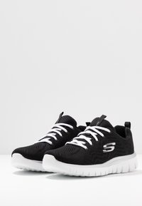 Baskets de sport noires avec un dessus en tricot, lacets blancs et semelle blanche rembourrée. Présente un logo sur le côté et une languette au talon.