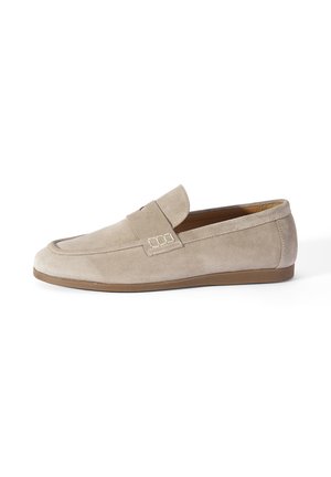 Beige suède loafer met een lage bruine zool en een decoratieve band over de wreef, zijaanzicht op een witte achtergrond.