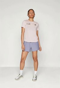 Ανοιχτό ροζ αθλητικό μπλουζάκι κοντό μανίκι με λογότυπο, συνδυασμένο με χαλαρές λεβάντες βερμούδες. Το μοντέλο φοράει λευκές κάλτσες και ασημένια αθλητικά παπούτσια.