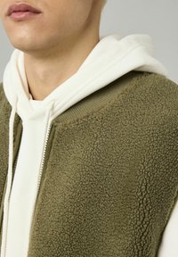 Gilet en polaire vert olive avec une surface texturée, fermeture éclair et encolure côtelée, superposé sur un sweat à capuche blanc avec des cordons de serrage.