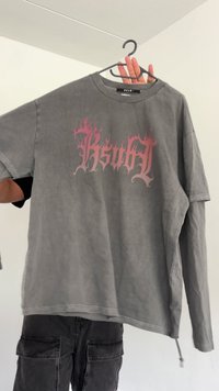 Person holds a gray long-sleeve shirt with pink flame-style text "KSUBI" on a black hanger against a white wall background.